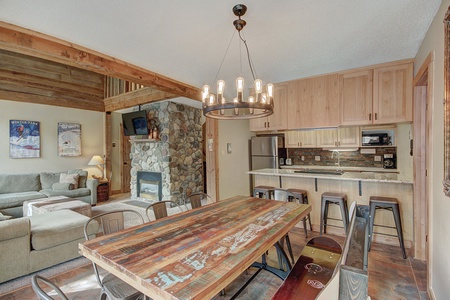 A rustic living and dining area with a wooden table, metal chairs, stone fireplace, wall paintings, and an open kitchen with wooden cabinets, stainless steel appliances, and a counter with stools.