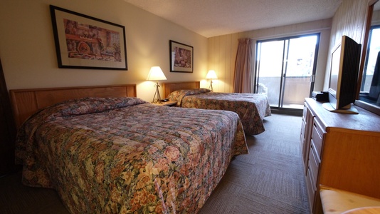 A hotel room with two beds, floral bedspreads, two bedside lamps, a flat-screen TV on a dresser, and a sliding glass door leading to a balcony.