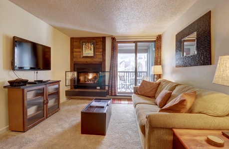 Cozy living room with a beige couch, wall-mounted TV, fireplace, and a coffee table. Sliding glass doors with a view of a balcony and outdoor scenery. Warm lighting from a lamp and wall mirror.