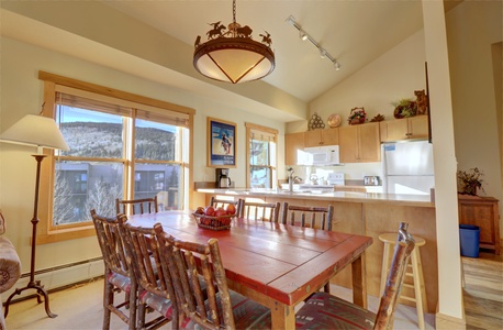 A well-lit kitchen and dining area with a rustic wooden dining table, chairs, a round chandelier, and large windows. The kitchen has wooden cabinets, white appliances, and various decorations.