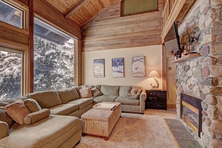 A cozy living room with a large sectional sofa, a stone fireplace, wall decor with posters, and large windows showing a snowy landscape outside.