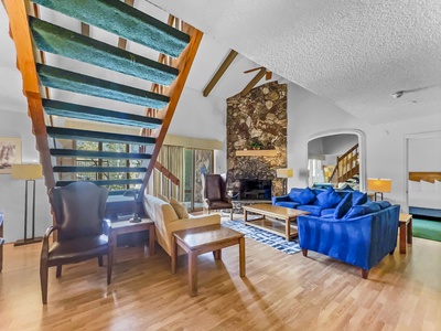 A living room with wooden floors, a stone fireplace, blue couch, armchairs, and a wooden staircase with green carpet leading to a loft. Large windows let in natural light.