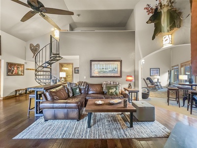 A cozy living room features a brown leather sectional, a wooden coffee table on a patterned rug, a ceiling fan, and a spiral staircase. The room is decorated with framed art and mounted deer heads on the wall.