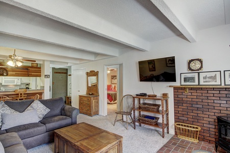 A cozy living room with a sofa, wooden coffee table, and TV mounted above a brick fireplace. The room has exposed beams, and an open doorway leads to a kitchen and a bedroom.