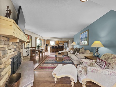 A cozy living room with a stone fireplace, TV, two sofas, an armchair, and a patterned rug. The room leads into an open kitchen and dining area with wooden cabinets and a table with four chairs.