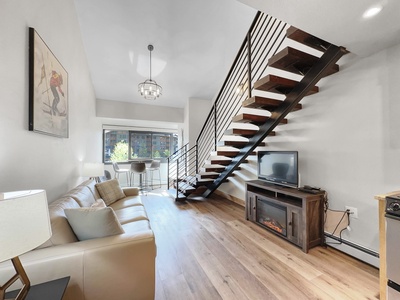 Modern loft apartment living room with a beige sofa, wall art, wooden floors, a TV on a console, and an open staircase leading to an upper level. Large windows allow natural light to fill the space.