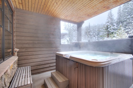 Steam rises from a warm, bubbling hot tub located on a covered patio, surrounded by snow-covered trees and a wintry landscape. Wooden steps lead up to the tub, and a bench sits nearby.