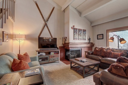 Living room with wooden furniture, a fireplace, TV, large windows, skis mounted on the wall, and a sofa set with cushions. A standing lamp is lit near the sofa.