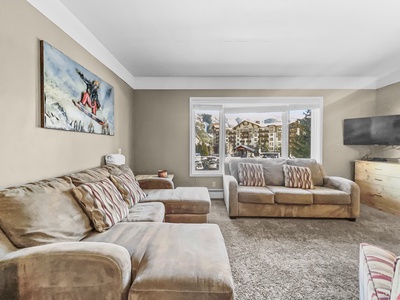 A living room with two beige sofas, a cushioned ottoman, a wall-mounted painting of a snowboarder, a large window showing exterior buildings, and a flat-screen TV on a wooden cabinet.