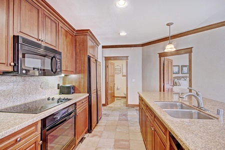 A modern kitchen featuring granite countertops, wooden cabinets, a built-in microwave, an electric stove, a double sink, and a hallway leading to another room. A toaster and knife set are on the counter.