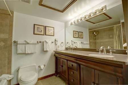 A bathroom with a large mirror, a double sink vanity, a toilet, and a towel rack with several folded towels. The walls are decorated with framed pictures.