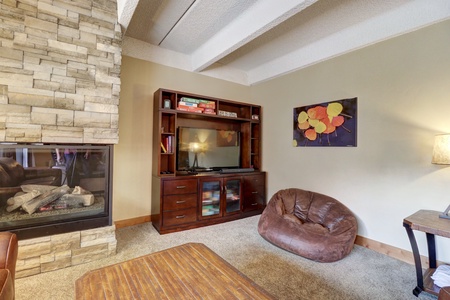 A living room featuring a stone fireplace, a wooden entertainment center with a TV and books, a brown bean bag chair, a lamp, and a wooden table. A painting with autumn leaves hangs on the wall.