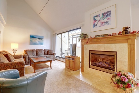 A cozy living room with a fireplace, floral arrangement, and comfortable seating. There is a painting on the wall and sliding glass doors leading to a balcony.