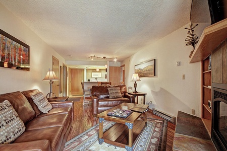 A cozy living room with leather sofas, a coffee table, and lamps, leading into a kitchen area. A painting and a framed picture adorn the walls.