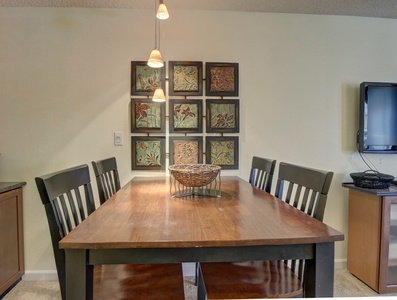 A dining table with four chairs, a centerpiece basket, wall art comprising nine square pieces, pendant lights above, and a wall-mounted TV in a cozy room.