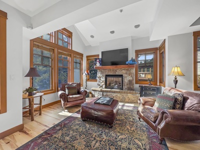 A cozy living room with large windows, a stone fireplace, leather furniture, and a patterned rug on wooden flooring. A flat-screen TV is mounted above the fireplace.