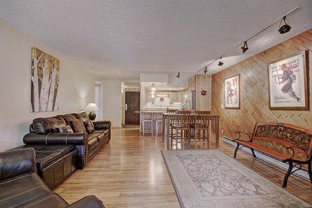 Living room with wooden floors, two leather couches, a wall-mounted poster, a dining table with chairs, and kitchen in the background.