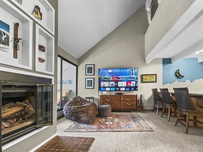 A living room with a wall-mounted TV, a fireplace, a beanbag, a patterned rug, and a bar area with high chairs. The walls have assorted artworks, and there's a sliding glass door to an outdoor space.