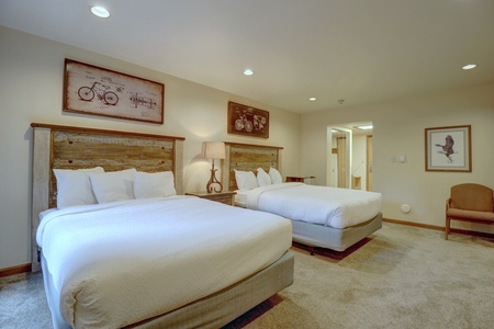 A hotel room with two queen-sized beds, wooden headboards, nightstand with a lamp, framed artwork on the walls, carpeted floor, and a door leading to another room.