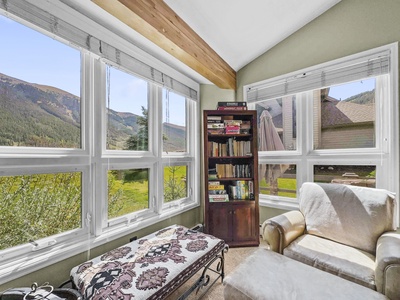 A cozy sunroom with large windows offering a mountain view. The room features a cushioned armchair, a patterned ottoman, and a bookshelf filled with books.