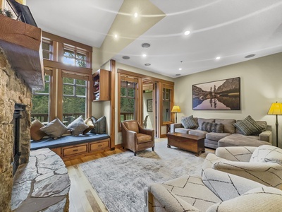 A cozy living room with a stone fireplace, built-in window seating, a gray couch, armchairs, a coffee table, and a large wall painting. Light streams in from large windows providing a view of trees.