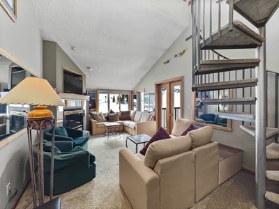 A cozy living room with beige sofas, green chairs, a fireplace, a TV, and a spiral staircase. Large windows and a door provide natural light and a view outside.