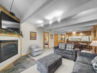 A living room with a fireplace, mounted TV, gray couch, armchair, and an ottoman. The room leads to a kitchen and a bedroom. Walls feature wood paneling and paintings. Ceiling has exposed beams and lights.