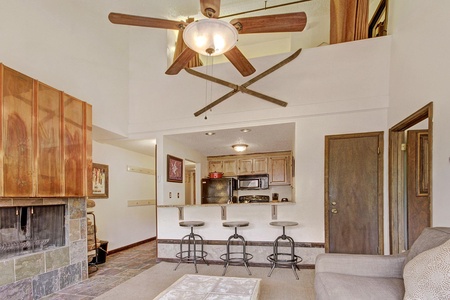 A cozy living area with a ceiling fan, fireplace, and a small kitchen. The kitchen has a breakfast bar with three stools. The space features wood accents and warm lighting.