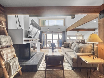 A cozy living room with wooden beams, a fireplace, a wall-mounted TV, a beige sofa, and a coffee table. Large windows and a sliding door lead to a balcony with outdoor seating.