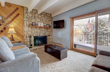 A living room with a stone fireplace, grey armchair, and a wall-mounted TV. There's a wooden coffee table, a sliding glass door leading to a balcony with a snowy view, and ski decor on the wall.