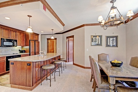 A kitchen and dining area featuring wooden cabinetry, a granite island with bar stools, a rectangular dining table with six chairs, and a chandelier. The space is decorated with framed pictures on the wall.