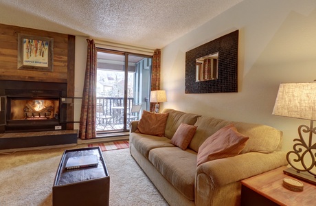 Cozy living room with a beige sofa, decorative pillows, coffee table, and a lit fireplace. Sliding glass door opens to a balcony with seating. Warm lighting from lamps creates an inviting atmosphere.