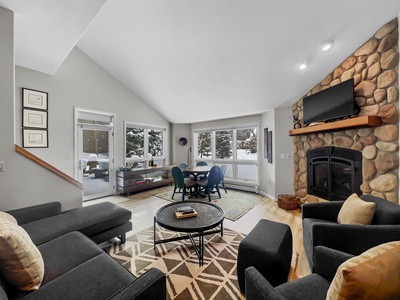 A modern living room features a stone fireplace with a TV above it, a round coffee table, two black armchairs, a sofa, and large windows overlooking a snowy landscape.