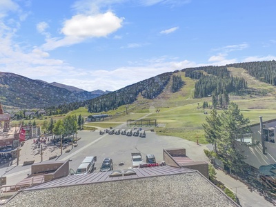 A mountainous landscape with grassy slopes, patches of trees, and a ski lift. In the foreground is a parking area with vehicles and a few buildings. The sky is blue with scattered clouds.