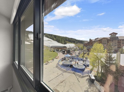 View through a window showing an outdoor recreational area with trampolines, people walking, and buildings in the background on a sunny day.