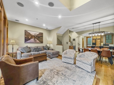 A cozy living room with a sectional sofa, two armchairs, a brown leather chair, a large area rug, and a dining area in the background featuring a long wooden table and chairs.