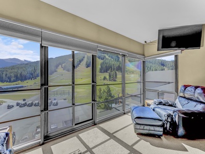 A room with large windows offering a scenic view of mountains and greenery, featuring a blue recliner, a TV mounted near the ceiling, and a carpeted floor.