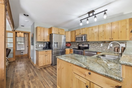 Modern kitchen with wooden cabinets, stainless steel appliances, and a granite countertop. The kitchen features a tile backsplash, overhead lighting, and hardwood floors. Doorway leads to another room.