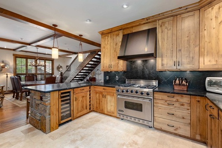 A spacious kitchen with wooden cabinets, black countertops, a large stainless steel stove, and a wine fridge. A staircase and dining area are visible in the background.