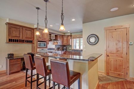 A modern kitchen with wooden cabinets, granite countertops, stainless steel appliances, a built-in desk, and a breakfast bar with three leather stools. Three pendant lights hang above the bar.