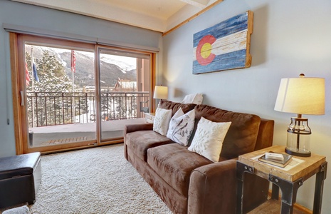 Living room with a brown sofa, two side tables with lamps, a Colorado flag wall art, and a sliding glass door leading to a balcony with a mountain view.