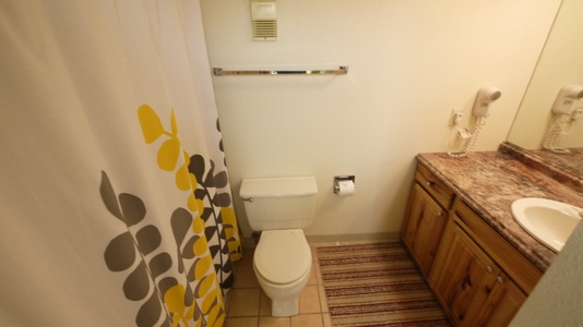 A bathroom with a white toilet, brown wooden vanity, and granite countertop. There is a gray and yellow shower curtain and a striped rug on the tiled floor.