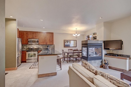 Open-plan living area featuring a kitchen with stainless steel appliances, a dining table, a sitting area with a beige sofa, and a wall-mounted TV next to a double-sided fireplace.