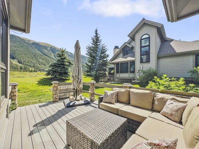 A patio with beige cushions and a wicker table overlooks a grassy area with pine trees and mountains in the background. An umbrella is centered on the patio. Two houses are visible on the right side.