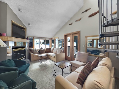 A spacious living room with beige and teal seating, a fireplace, large windows, and a spiral staircase. Walls are adorned with decorative wooden accents.