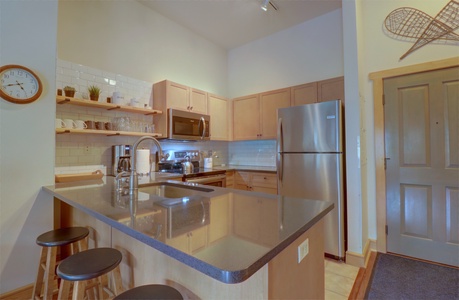 Modern kitchen with light wood cabinets, stainless steel appliances, and a dark countertop island with barstools. Open shelving holds kitchen accessories, and a clock hangs on the wall.