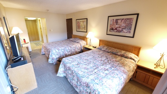 A hotel room with two beds covered in floral bedspreads, a nightstand with lamps, framed art on the walls, and a television. The room is well-lit with an open doorway leading to a hallway.