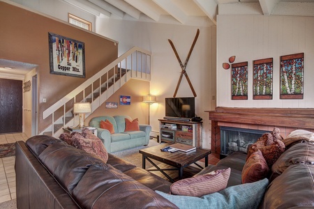 Living room with a staircase, a sofa, a loveseat, a coffee table, a flat-screen TV, and a fireplace. Wall decor includes crossed skis and art pieces. The room has a cozy, rustic feel.