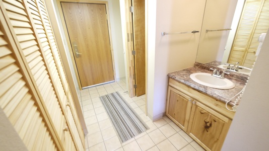 A small bathroom with a sink and vanity on the right, a closet with louvered doors on the left, and a wooden door at the end of the tiled floor hallway. .