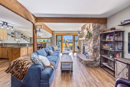 A spacious living room with a blue sofa, stone fireplace, and wooden coffee table. The room is connected to an open kitchen and features large windows with a view of the outdoors.